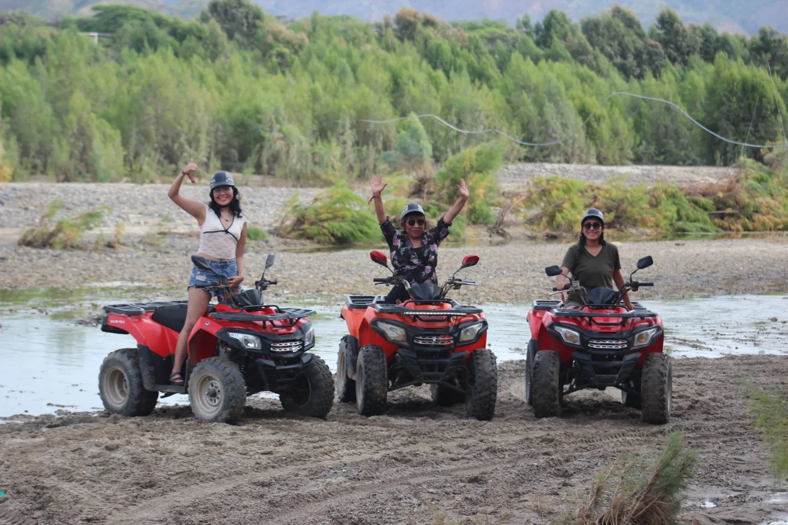 Tres personas con casco y los brazos en alto posan con sus cuatrimotos rojas en la orilla de un río poco profundo