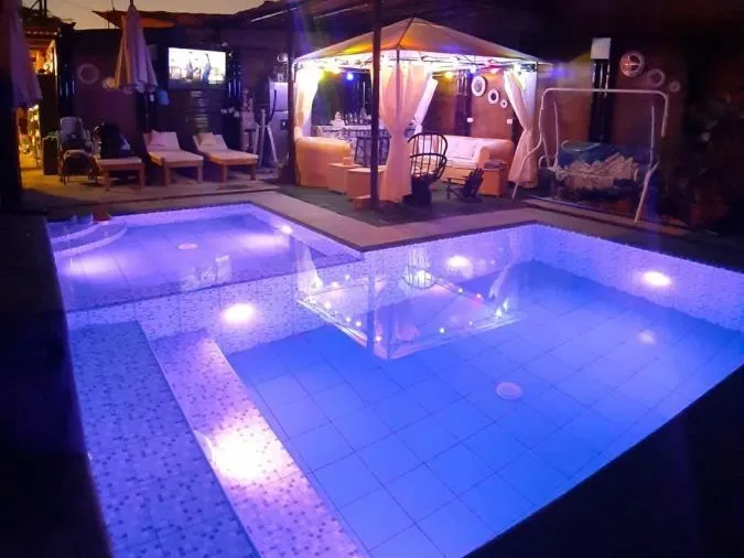 Piscina iluminada de noche, vacía, con una carpa de cortinas blancas, un living y un columpio al fondo