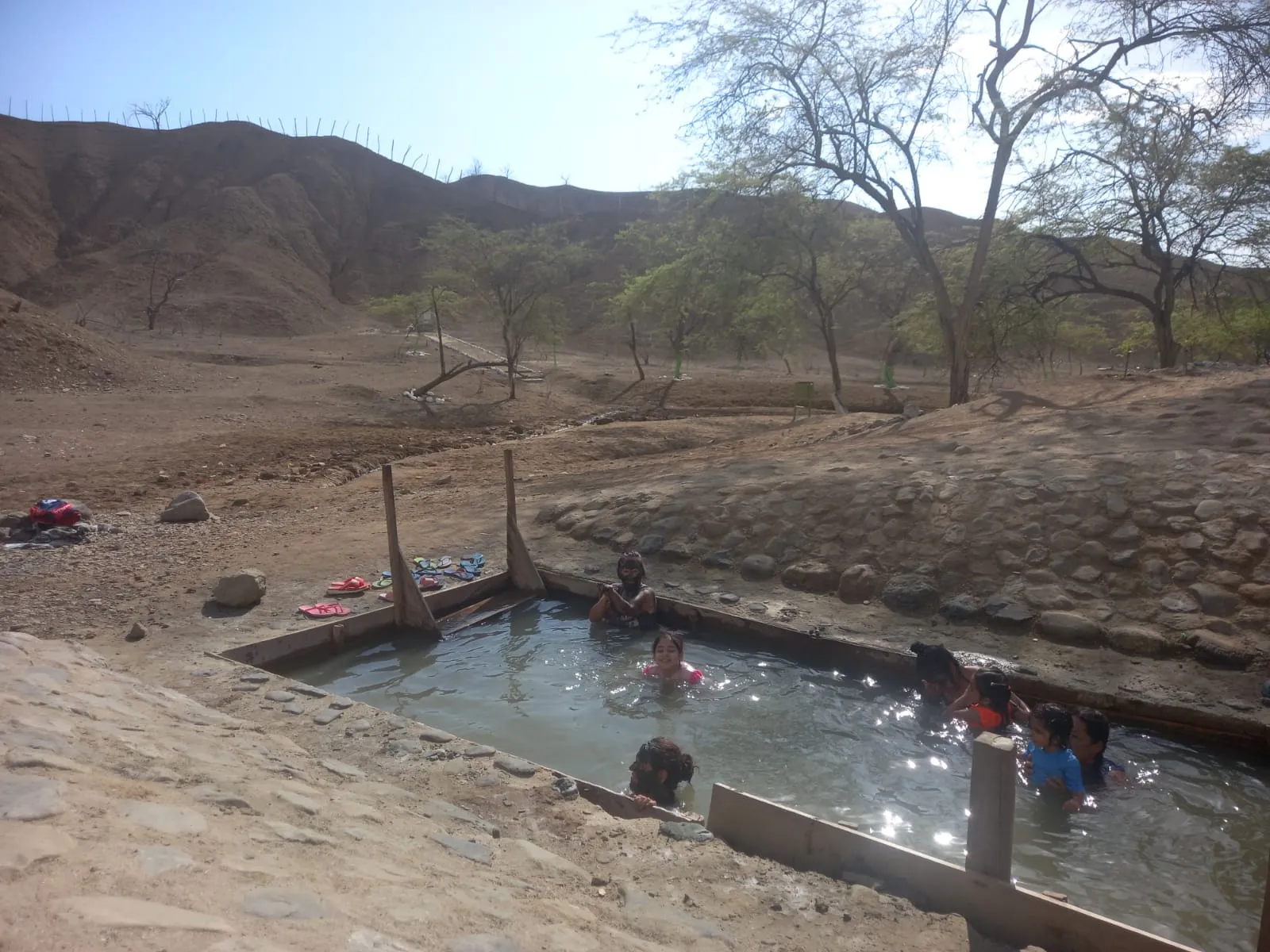 Varias personas se bañan en una poza con bordes de madera y piedra, en un valle seco con cerros y árboles
