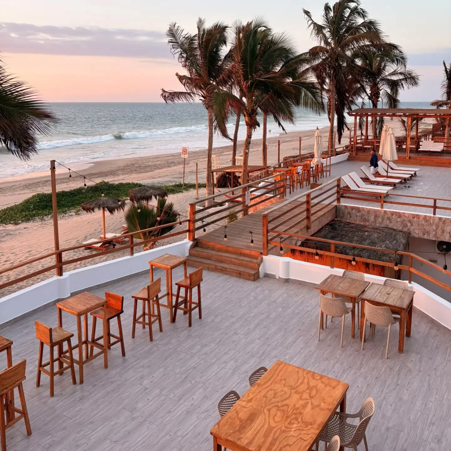 Terraza de madera con mesas y taburetes frente a la playa, con palmeras y el mar al atardecer