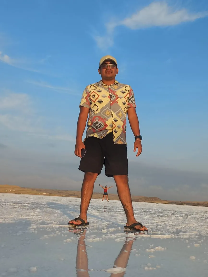 The legs and feet of a person standing on the white, reflective floor of the salt flat, with another person in the distance raising their arms and the horizon behind