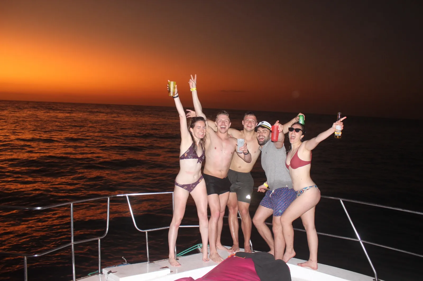 Grupo de personas brindando y levantando los brazos en la cubierta de un yate, bajo un cielo naranja de atardecer sobre el mar